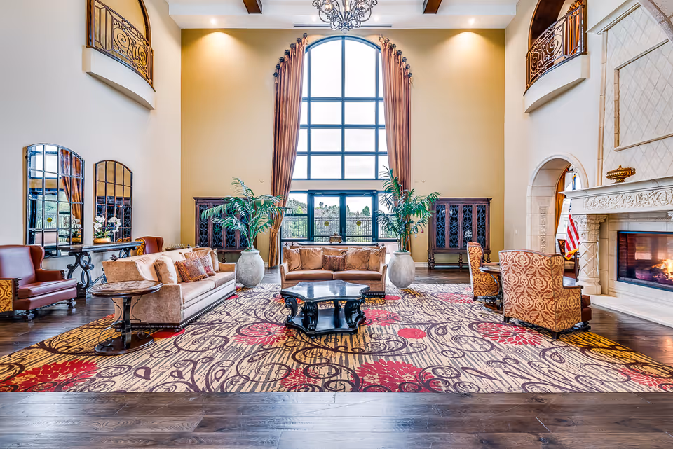 Spacious elegant lounge with high arched window, sofas and armchairs arranged around a central table, potted plants and a fireplace.