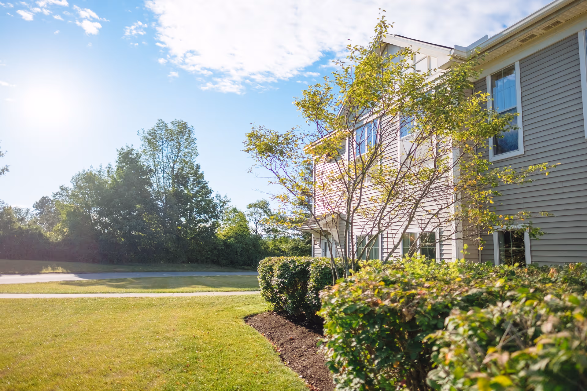 Exterior view of a two-story building with beige siding partially visible on the right side, surrounded by green bushes and trees under a bright blue sky with scattered clouds and sunlight.
