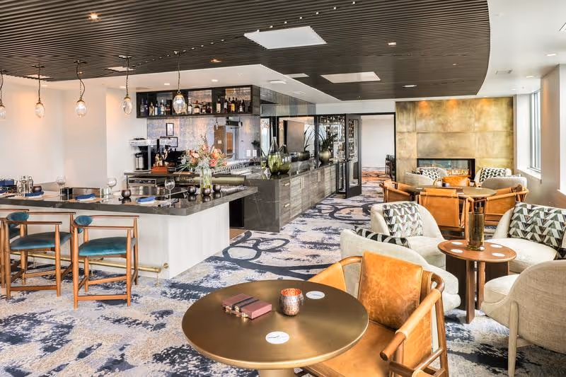 A modern, stylish lounge area featuring a bar with hanging pendant lights, a countertop with bar stools, and several seating arrangements including armchairs and small round tables. The room has a patterned carpet, a wooden slatted ceiling, and a fireplace with a golden panel backdrop. Large windows allow natural light to fill the space.