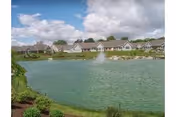 A pond with a central fountain in front of a row of cottage-style homes under a cloudy sky.