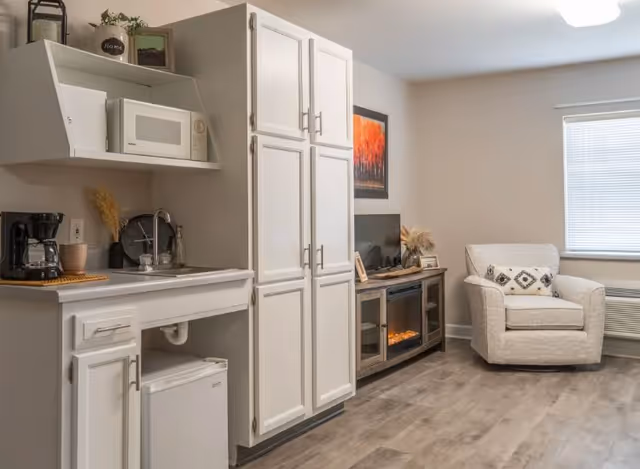 A cozy living area featuring a small kitchenette with white cabinets, a microwave, coffee maker, and mini refrigerator. Adjacent to the kitchenette is a wooden TV stand with an electric fireplace and a flat-screen TV. A comfortable armchair with decorative pillows sits near a window with closed blinds, and the room has light-colored walls and wood flooring.