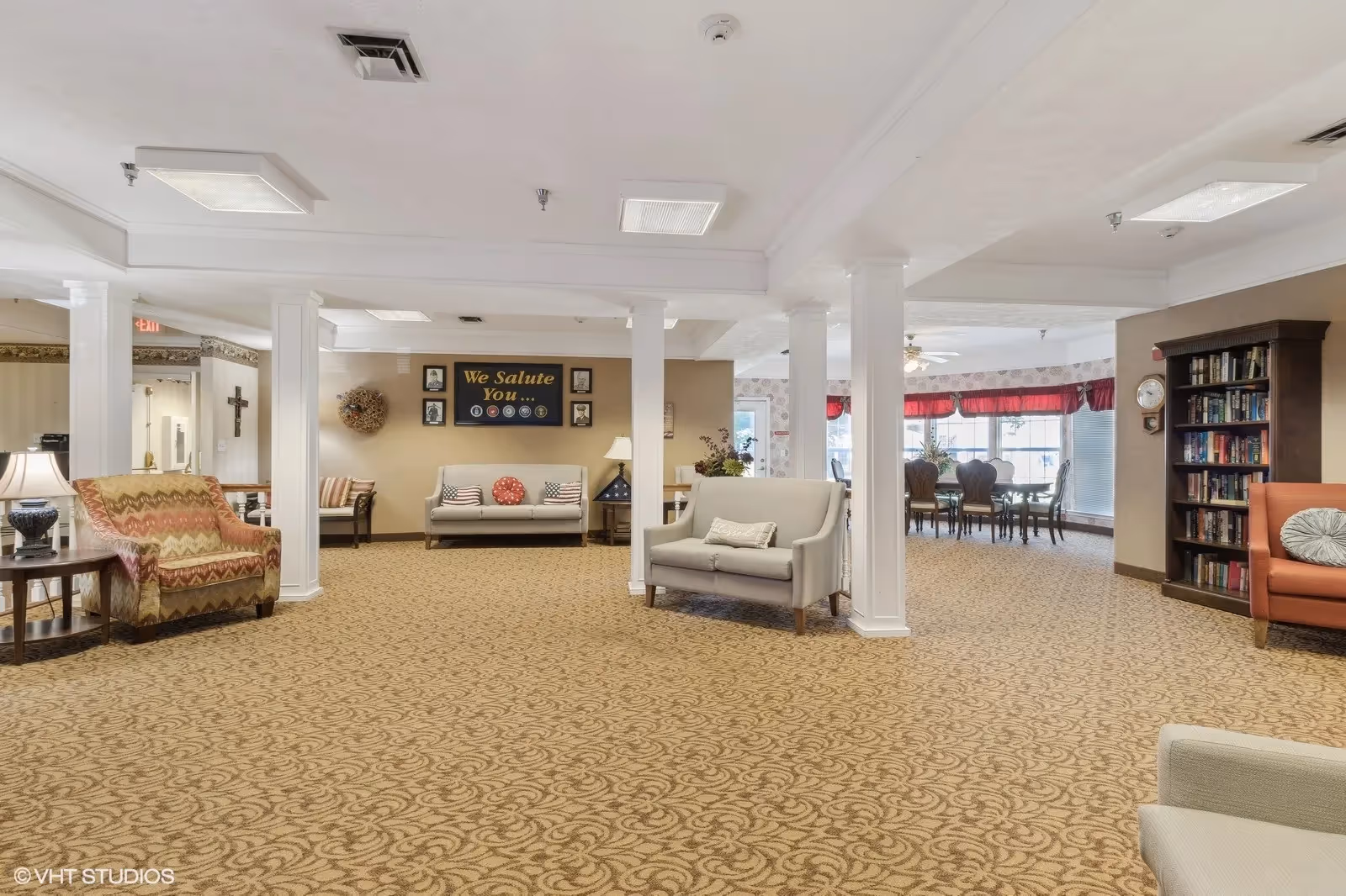 A spacious senior living facility common area with patterned carpet, multiple seating arrangements including armchairs and sofas, white columns, a bookshelf filled with books, and a dining area with a table and chairs near large windows with red valances. A wall display reads 'We Salute You' with military insignias and framed photos.