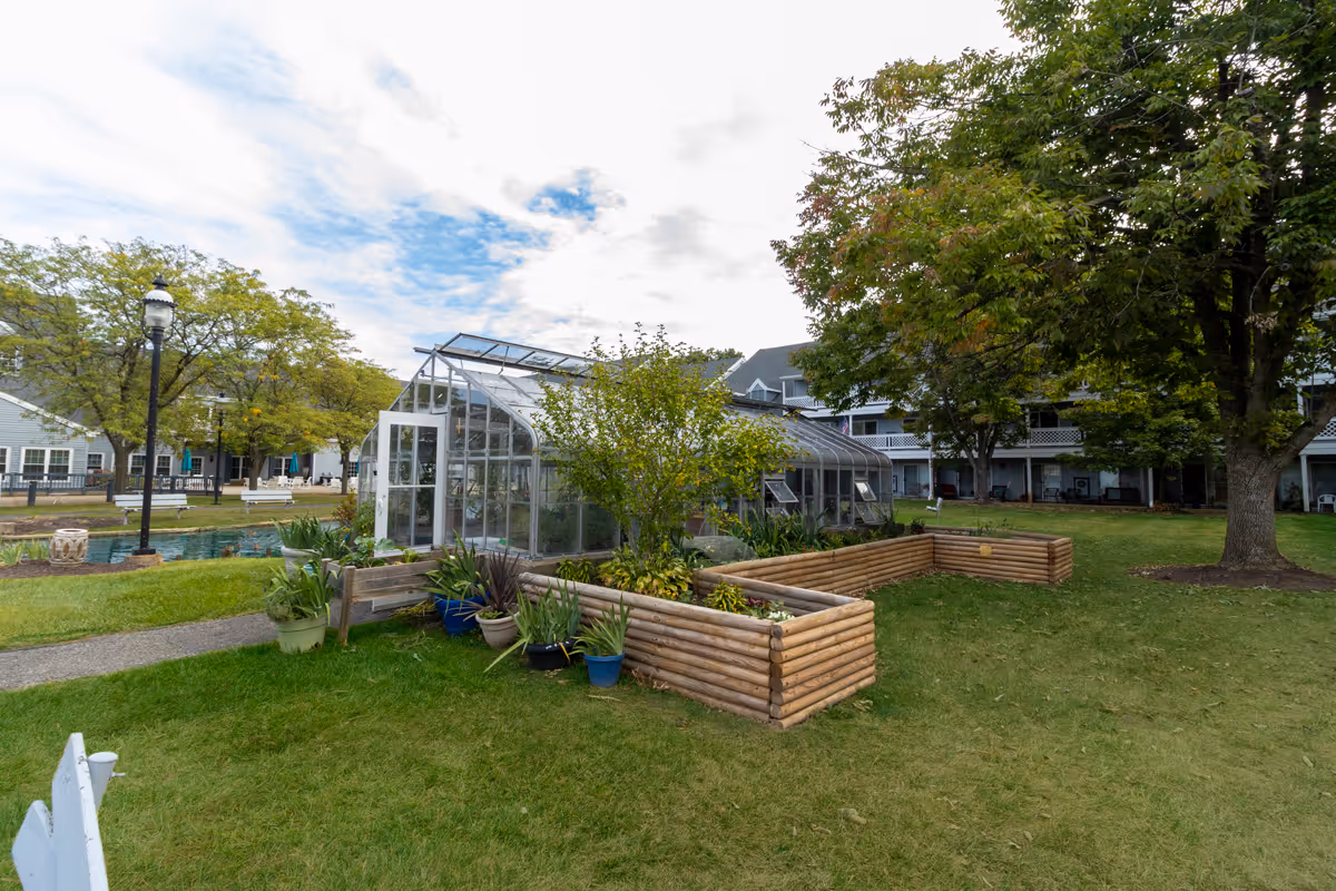A grassy courtyard with a glass greenhouse, raised wooden planters, trees and a multi-story residential building in the background.