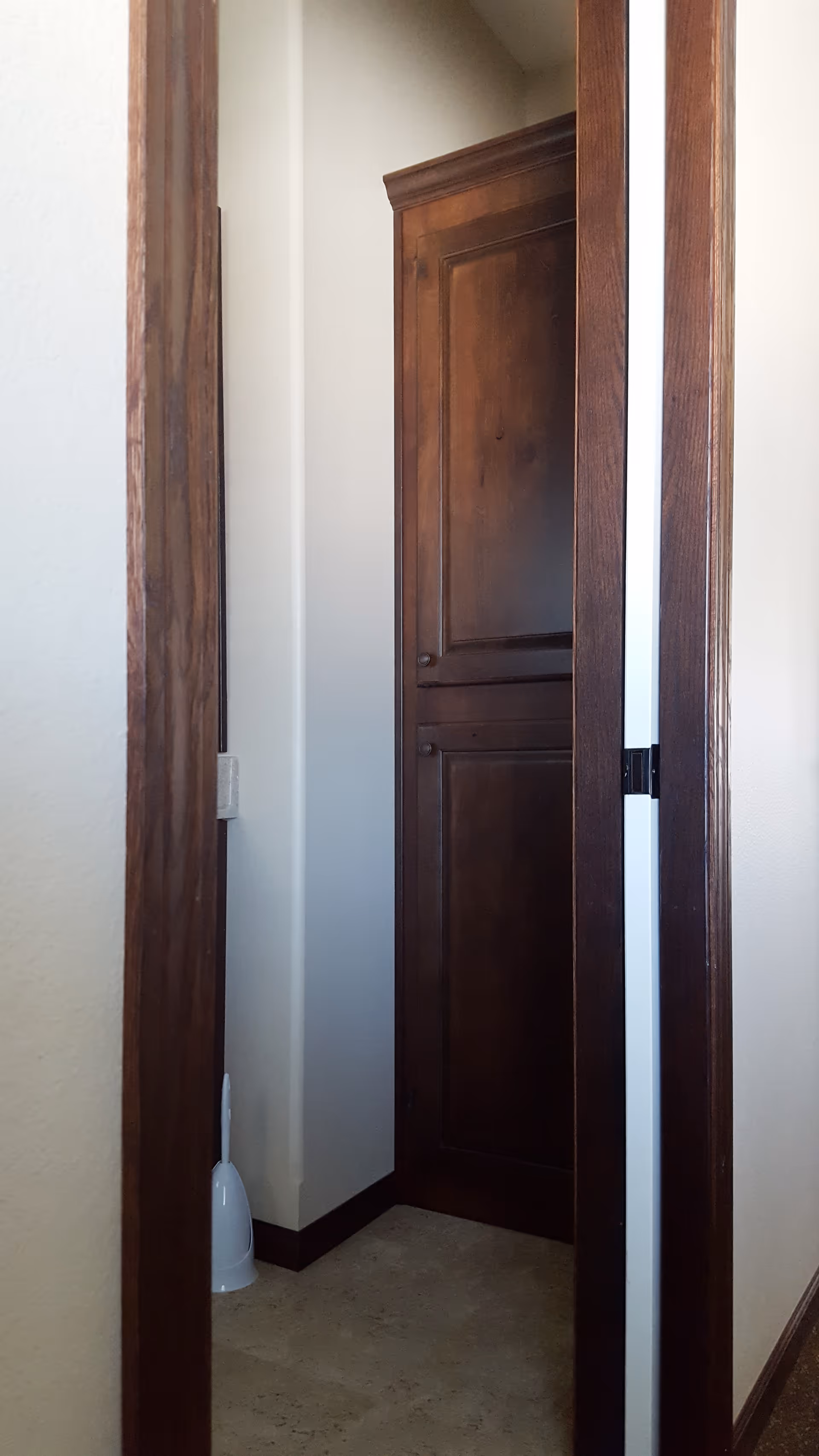 A narrow interior space with beige walls and a tiled floor, featuring a dark wooden cabinet with two doors and a white toilet brush holder placed on the floor in the corner.