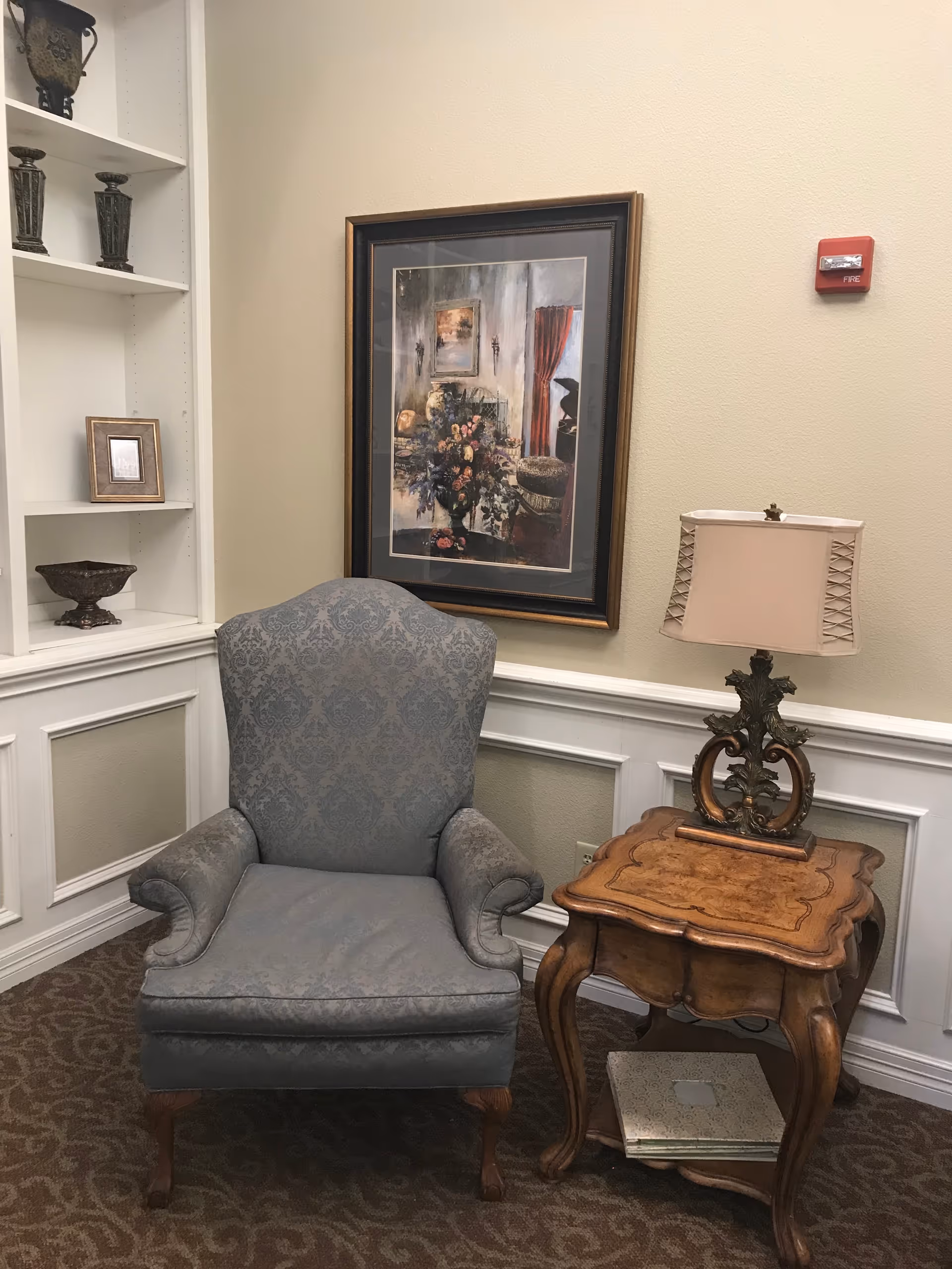 A cozy corner featuring a patterned blue armchair next to an ornate wooden side table with a decorative lamp. Behind the chair is a framed painting of a floral arrangement and a piano in a softly lit room. To the left, built-in white shelves hold decorative vases and a small framed picture. The walls are beige with white wainscoting, and the floor is carpeted with a subtle pattern.