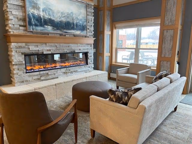 A cozy living room area featuring a modern stone fireplace with a long horizontal flame, a large painting above the fireplace, a beige sofa with patterned pillows, a brown armchair, a round brown ottoman, and a window showing an outdoor patio with chairs and tables.