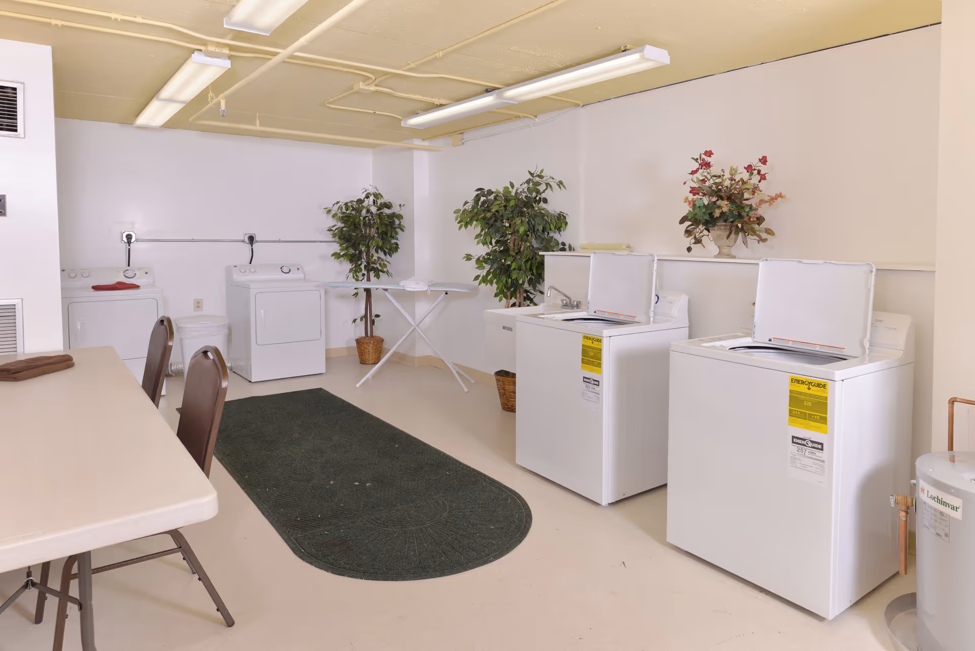 Laundry room with four white top-loading washing machines, two artificial plants in baskets, an ironing board, a folding table with two chairs, and a green rug on the floor. The ceiling is painted yellow and the walls are white.