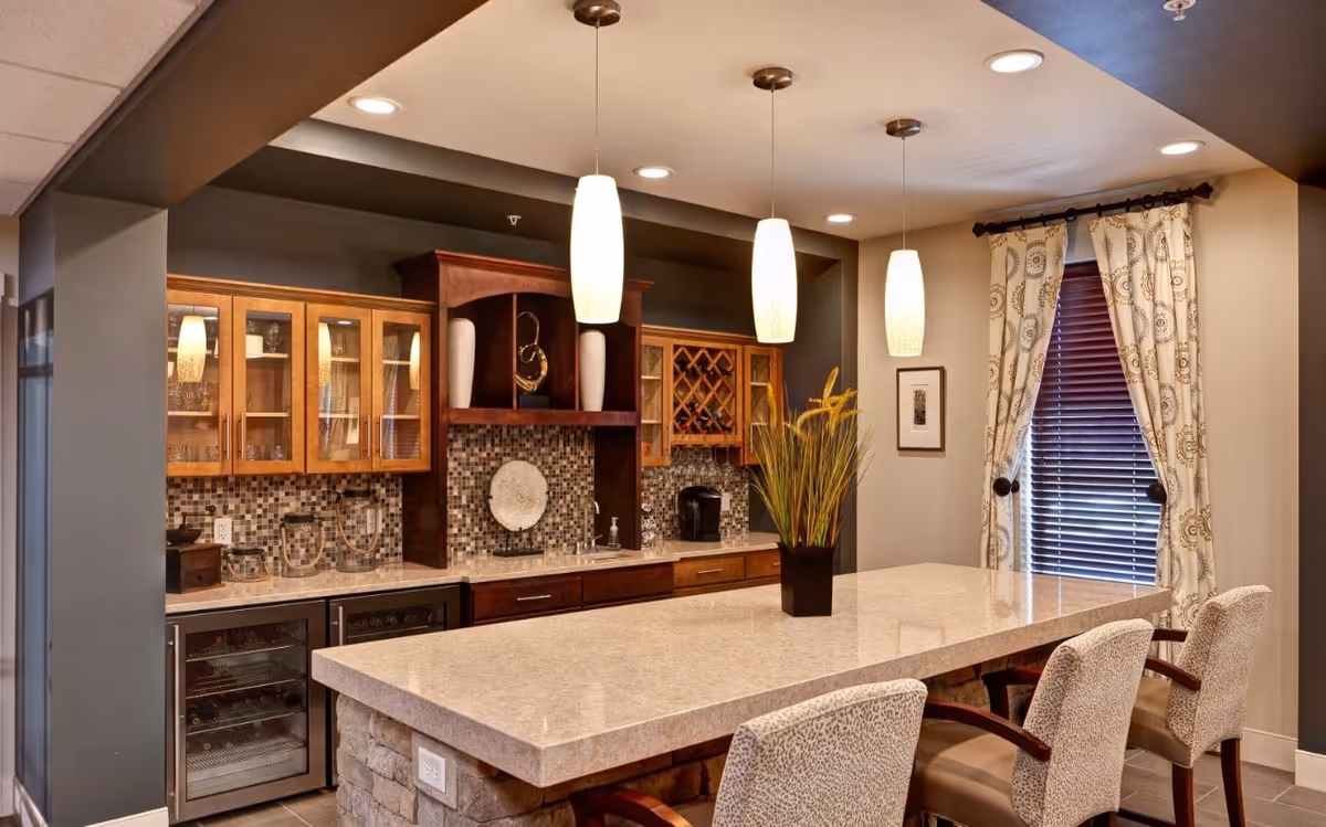 Large communal kitchen and bar area featuring a long stone island with seating, pendant lights, wood cabinetry, and a tiled backsplash.