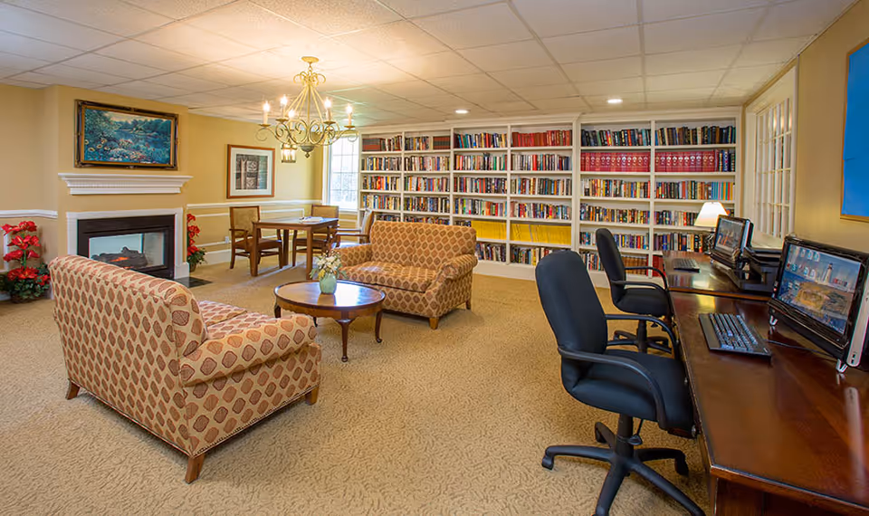 A cozy common area with patterned armchairs and a loveseat around a round wooden coffee table, a fireplace with a painting above it, a chandelier, a table with chairs near a window, a large bookshelf filled with books, and a desk with two computers and office chairs.