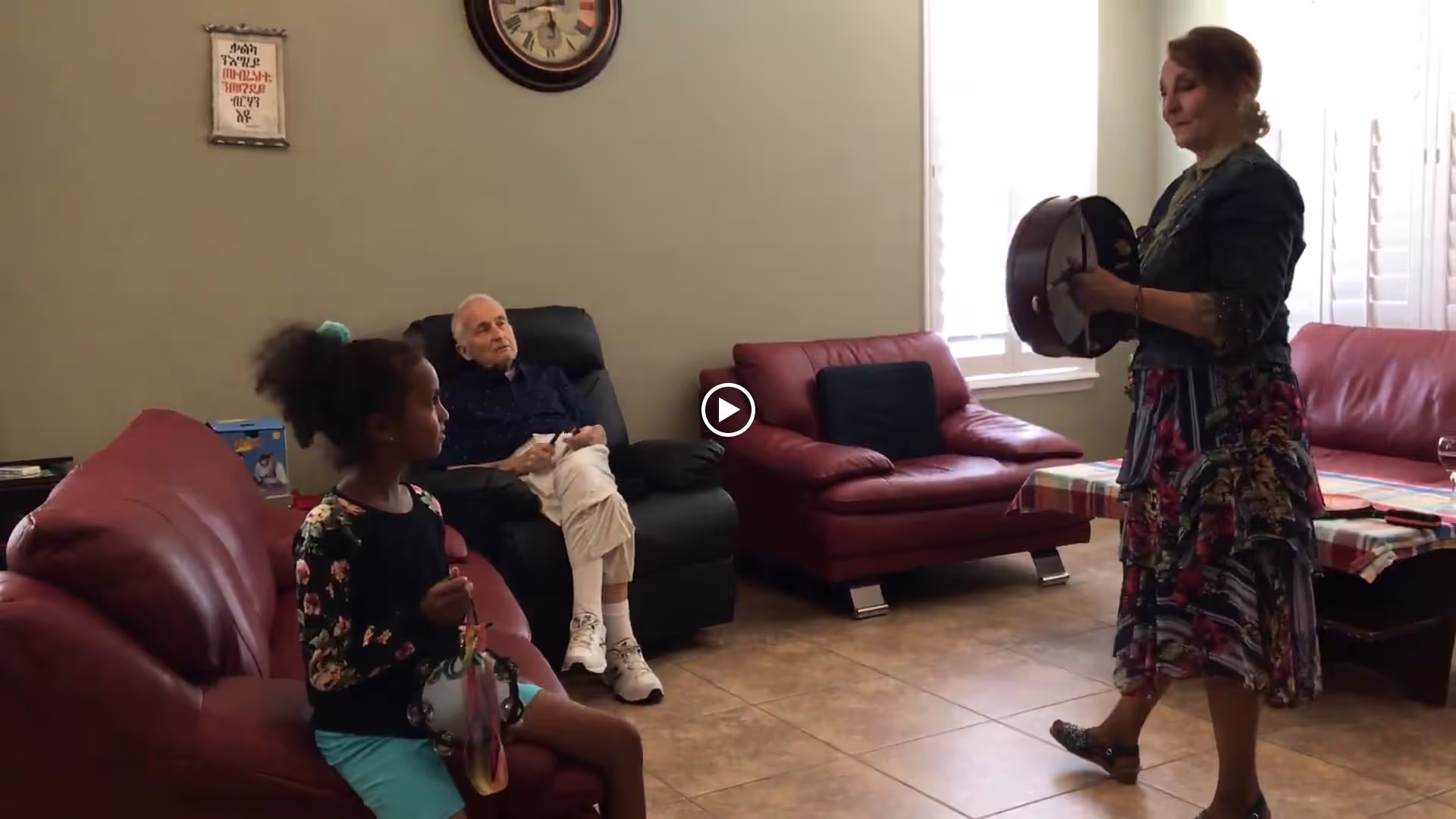 A living room with red leather couches and a recliner. An elderly man is sitting in the recliner, a young girl is sitting on one of the couches holding a tambourine, and a woman is standing playing a drum. The room has tiled flooring and a window with white blinds.