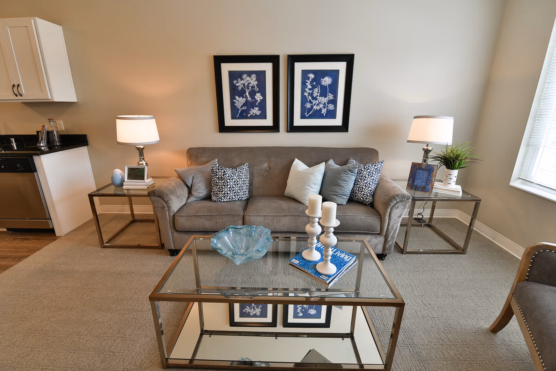 Neatly furnished living room with a gray sofa, glass coffee table, side tables with lamps, and framed botanical prints above the sofa.
