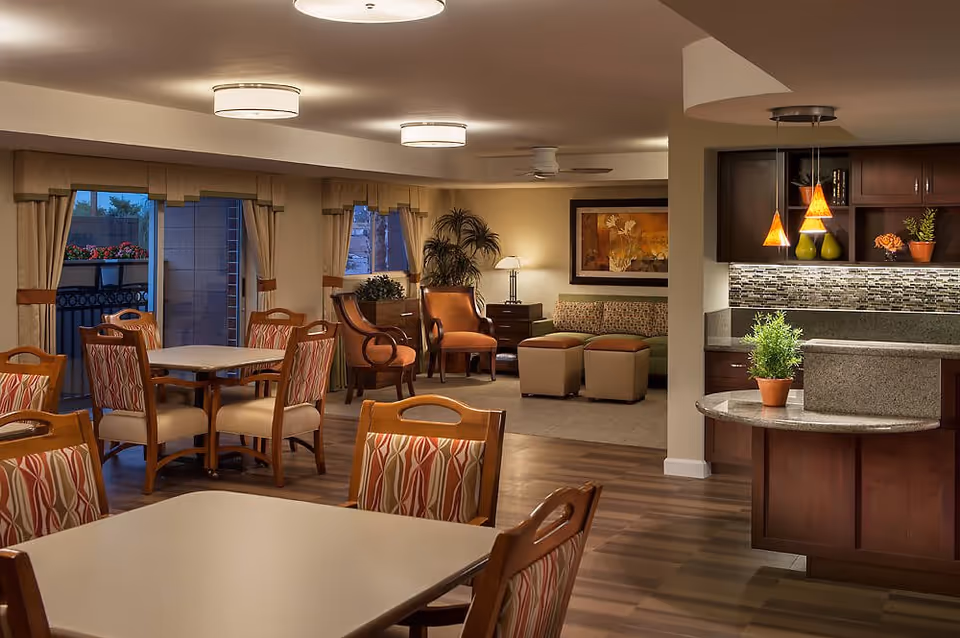 Interior view of a senior living facility showing a dining area with tables and chairs, a cozy living room with armchairs, a sofa, ottomans, plants, and a kitchen area with pendant lights and a granite countertop.