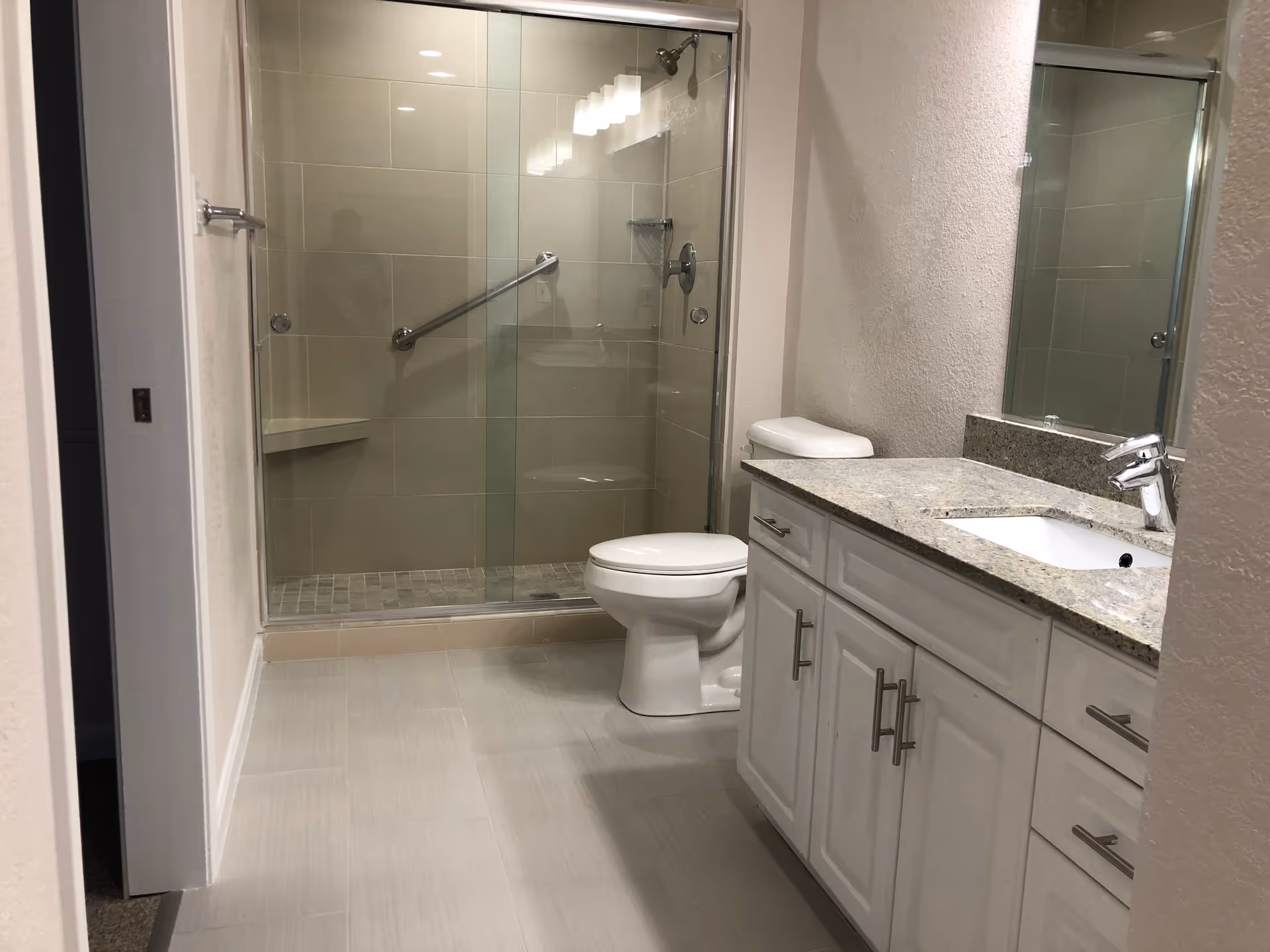 A clean and modern bathroom featuring a glass-enclosed shower with a grab bar, a white toilet, and a vanity with a granite countertop and a sink. The walls are light-colored, and the floor is tiled in a neutral tone.