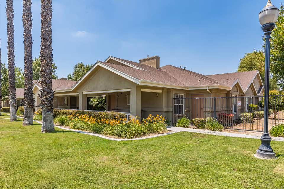 Exterior view of a single-story cottage-style senior living building with a covered porch, landscaped lawn, and palm trees.