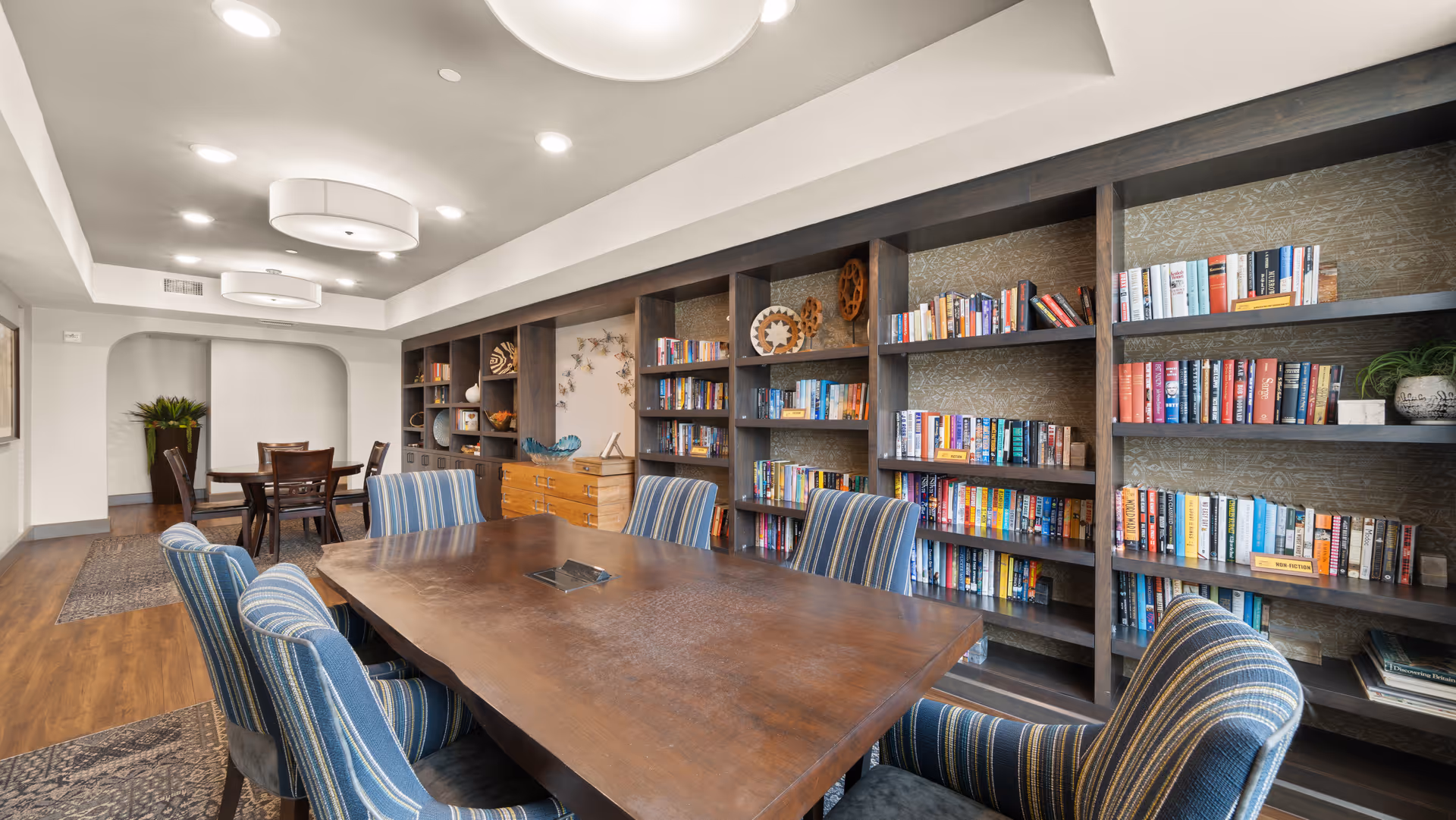 A well-lit interior room with a large wooden table surrounded by six blue-striped upholstered chairs. Behind the table is a wall with built-in dark wood bookshelves filled with books and decorative items. In the background, there is another smaller table with chairs and a plant in a corner. The room has wooden flooring and modern ceiling lights.