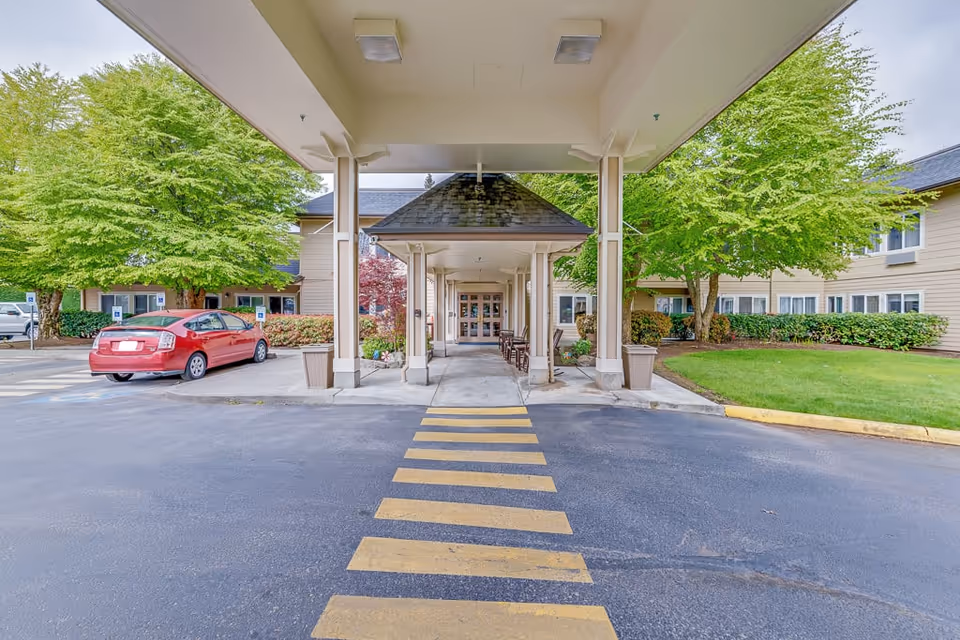 Entrance to a senior living facility with a covered drop-off area, a yellow pedestrian crossing on the asphalt, a red car parked to the left, green trees and shrubs surrounding the building, and a clear view of the double glass doors at the front.