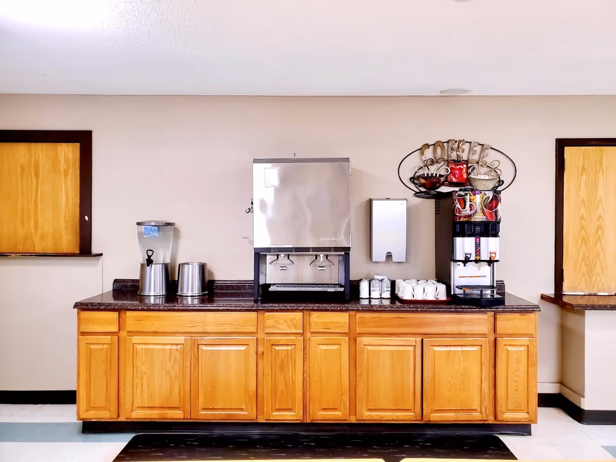 A beverage station with a water dispenser, ice machine, coffee machine, and several white coffee mugs on a wooden cabinet countertop in a room with beige walls and two wooden windows with dark brown trim.