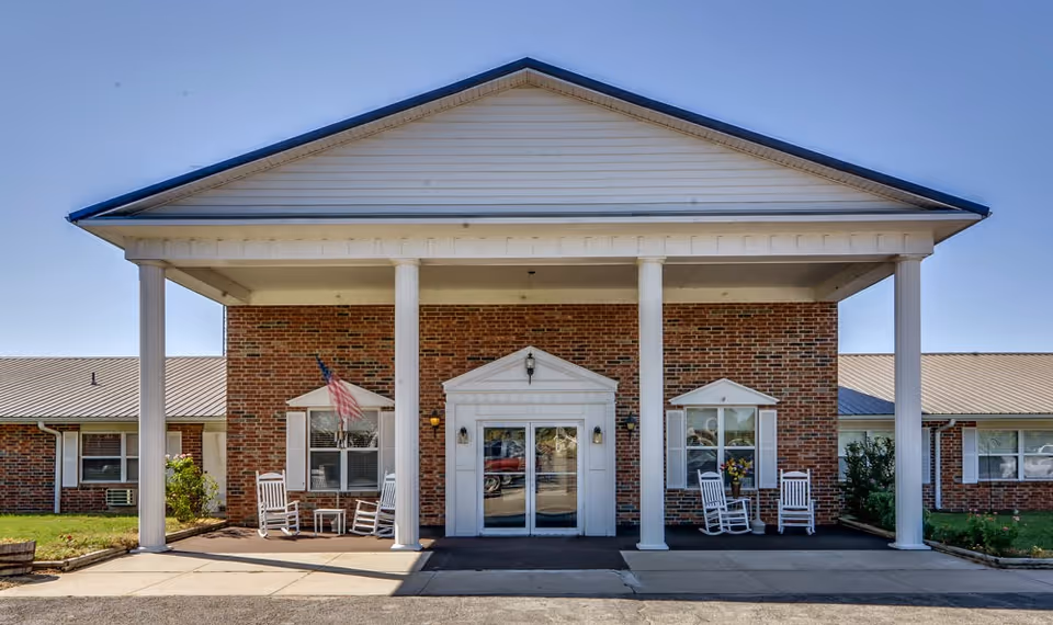 Front entrance of a brick building with a covered portico supported by white columns, rocking chairs on the porch, and an American flag.