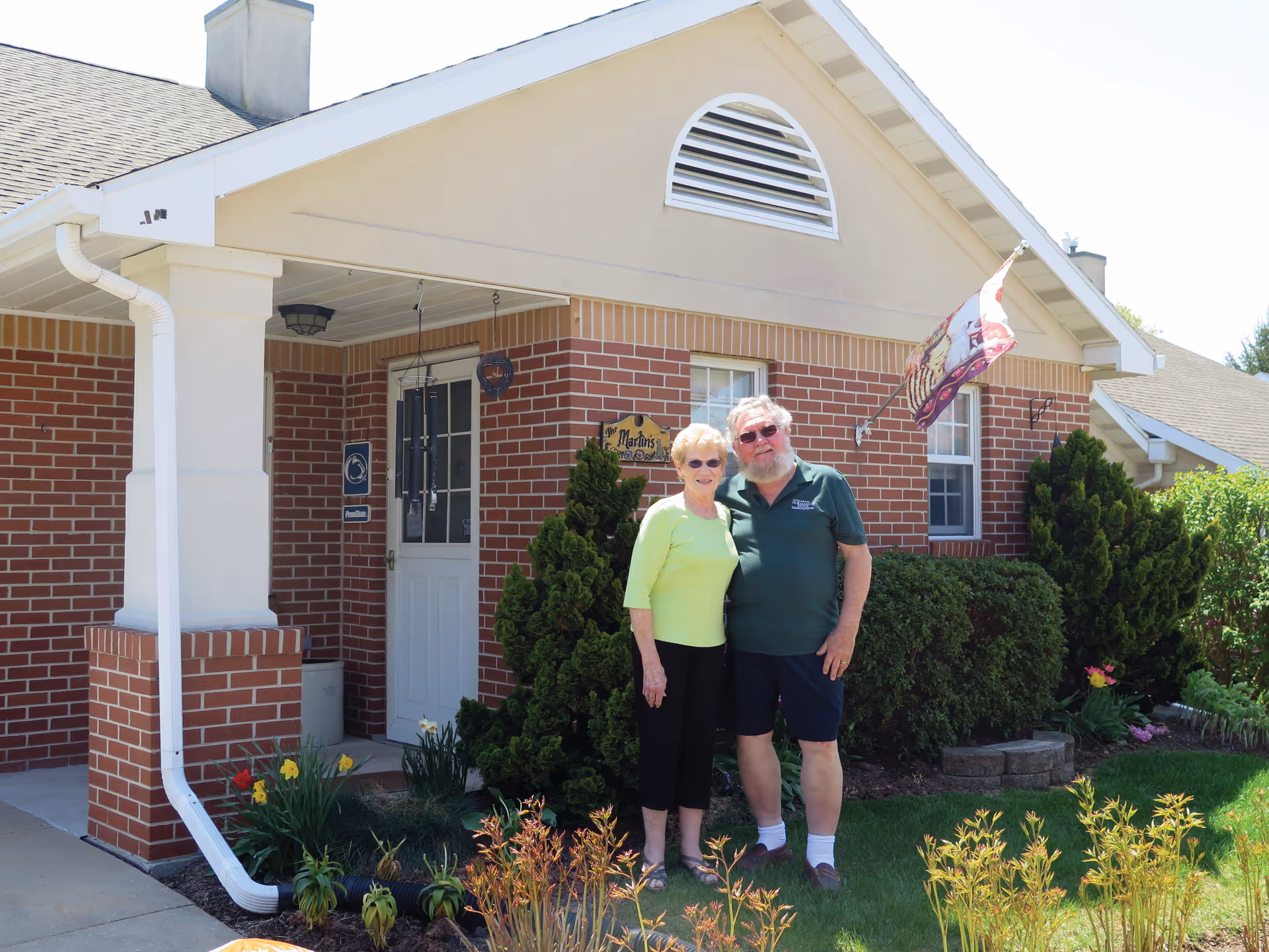 An elderly couple standing outside a brick house with a white door and a small garden with shrubs and flowers. The house has a beige upper facade and a flag mounted near a window. The couple is smiling and posing for the photo.