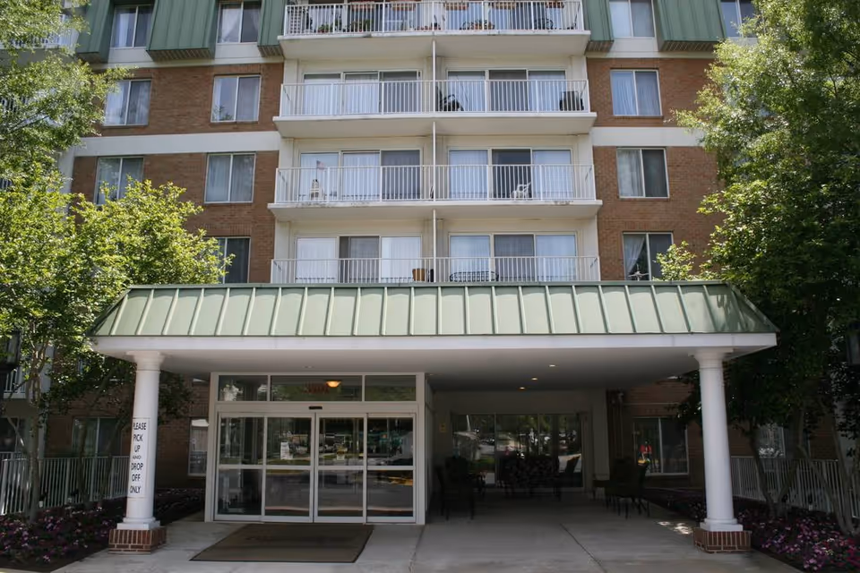Front entrance of a multi-story residential building with balconies, a green metal awning supported by white columns, and glass sliding doors. There are trees and flower beds on either side of the entrance.