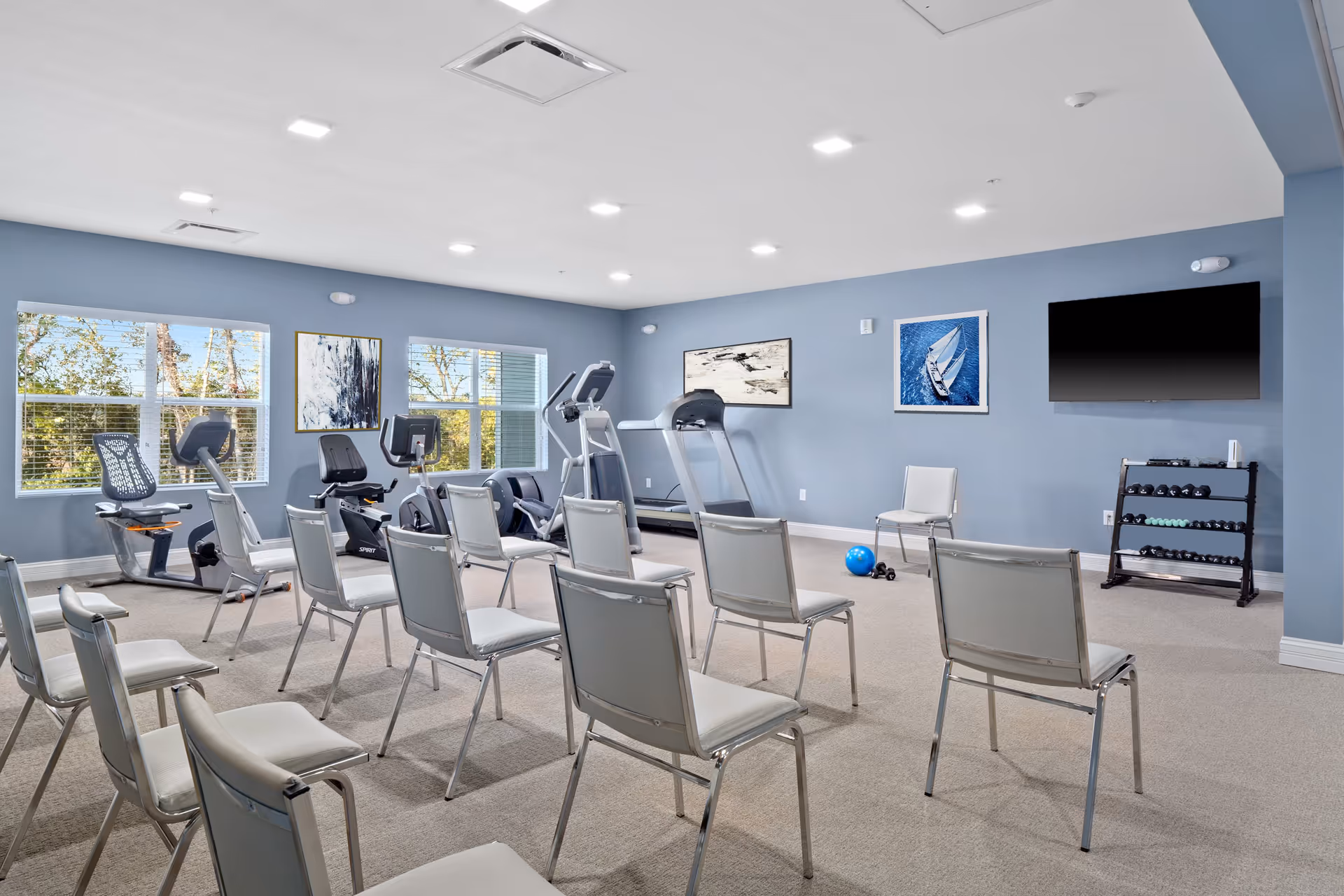 A fitness room with several exercise machines including stationary bikes and a treadmill, arranged behind rows of white chairs. The room has light blue walls, two large windows with views of trees outside, framed artwork on the walls, a flat-screen TV mounted on the wall, and a rack of dumbbells. The floor is carpeted and the ceiling has recessed lighting.