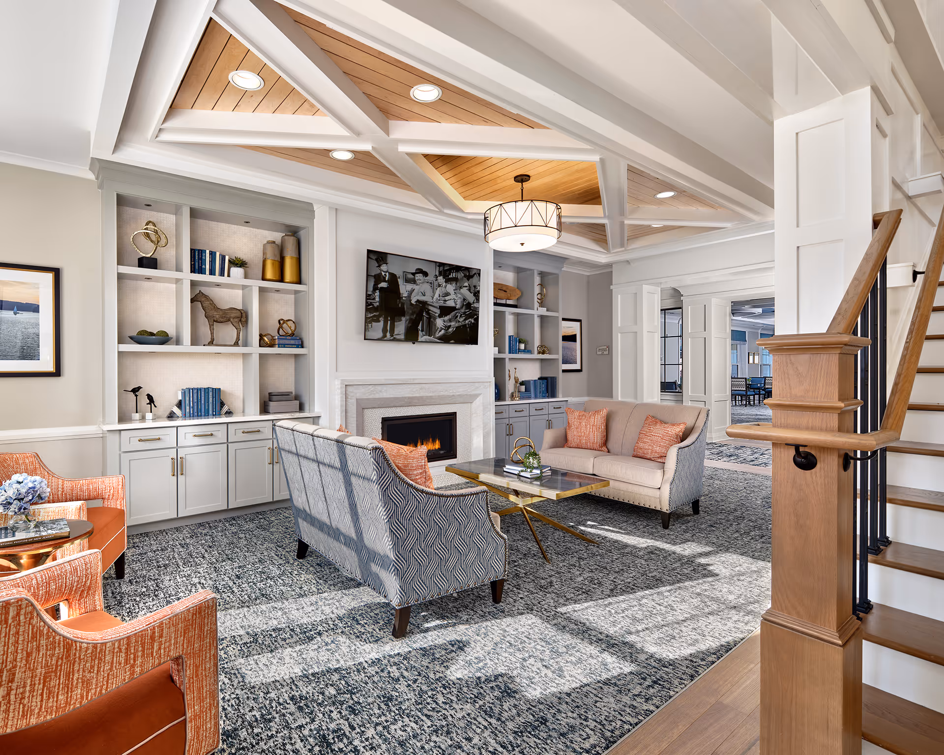 A bright and elegant living room area in a senior living facility featuring a patterned carpet, two upholstered sofas with orange accent pillows, two orange armchairs, a glass coffee table, built-in shelves with decorative items, a fireplace with a black and white photo above it, and a wooden staircase with black railings. The ceiling has a unique wooden panel design with recessed lighting and a central hanging light fixture.