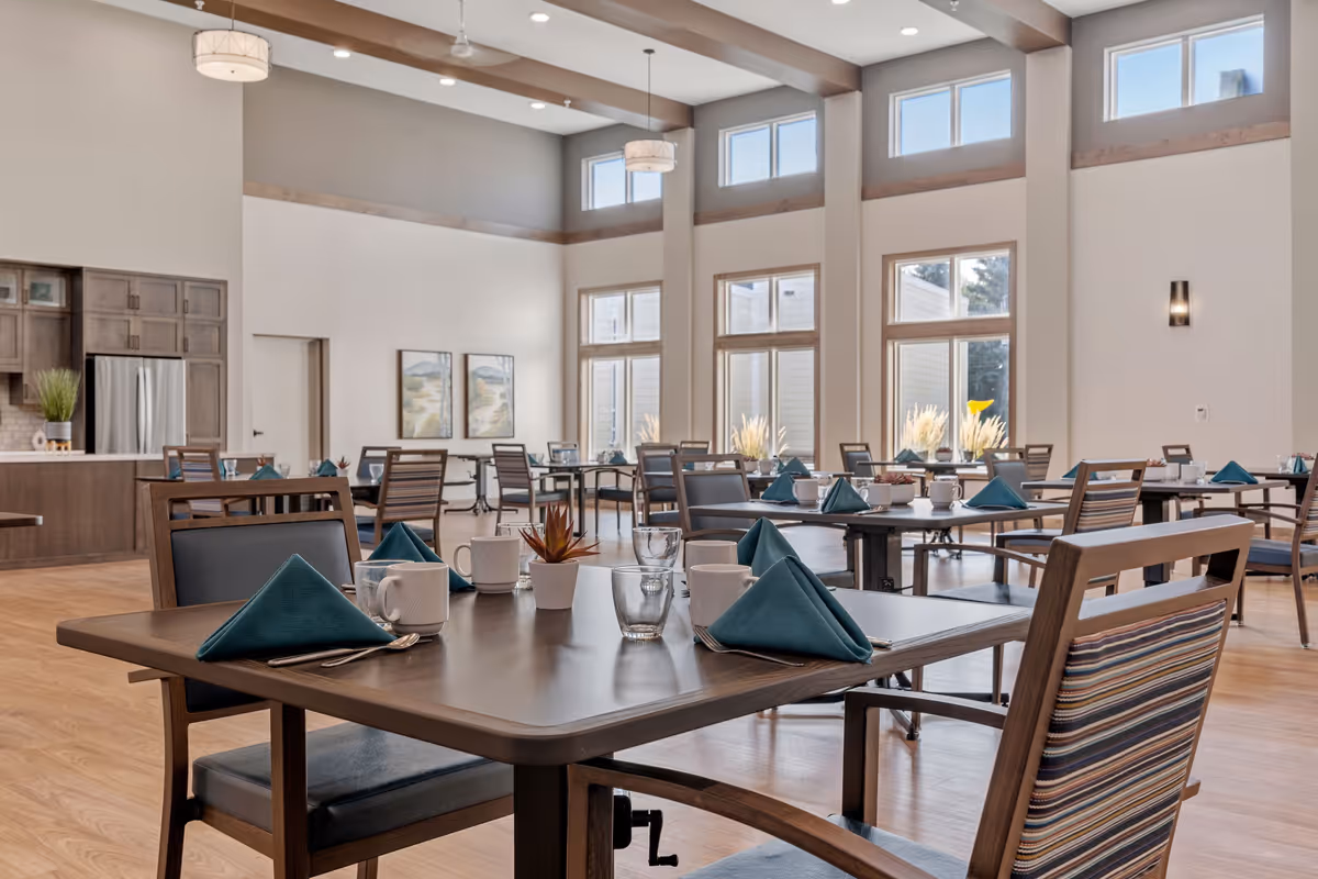 A spacious, well-lit dining room with multiple tables set with teal napkins, white mugs, glasses, and silverware. The room features large windows allowing natural light to fill the space, light wood flooring, and modern light fixtures hanging from the ceiling. There is a kitchen area with wooden cabinets and a stainless steel refrigerator in the background.