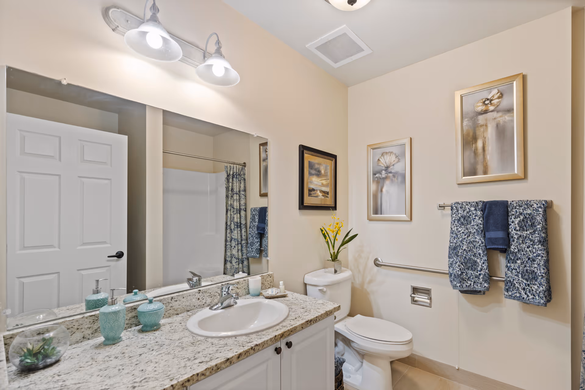 A clean and well-lit bathroom featuring a granite countertop with a white sink, decorative soap dispensers, and a small plant. There is a large mirror above the sink with two light fixtures. The bathroom includes a white toilet with a small plant on the tank, two framed pictures on the wall, and a towel rack holding two patterned blue towels and one solid blue towel. A shower with a patterned curtain is visible in the background.