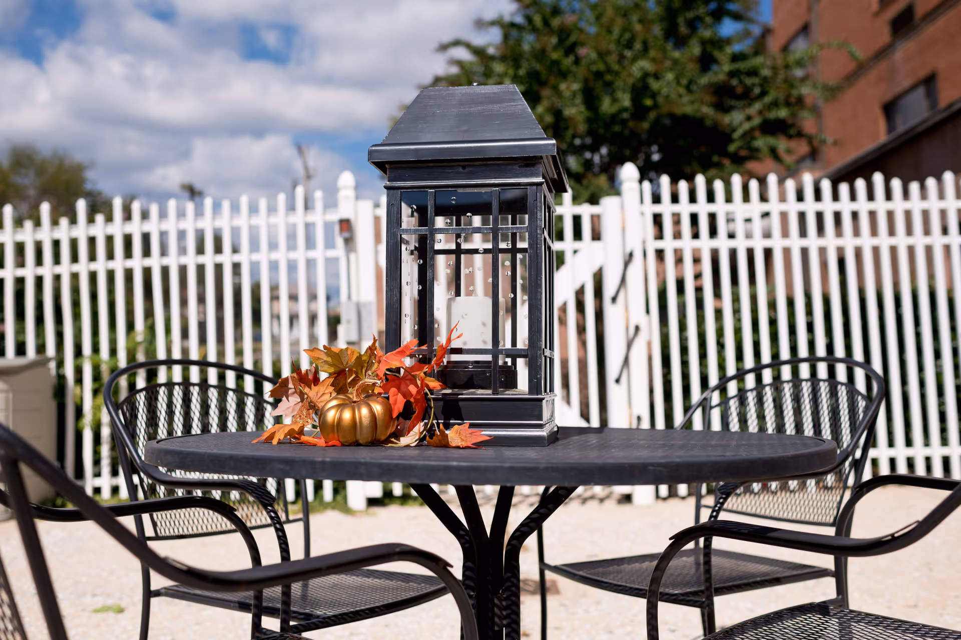 Outdoor patio area with a black metal table and four matching chairs. On the table is a decorative black lantern and a small golden pumpkin surrounded by autumn leaves. A white picket fence and some greenery are visible in the background under a partly cloudy sky.