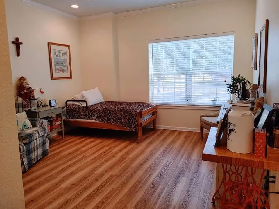 Simple bedroom with a single bed, armchair, side table, large window with blinds, and wood-effect flooring.