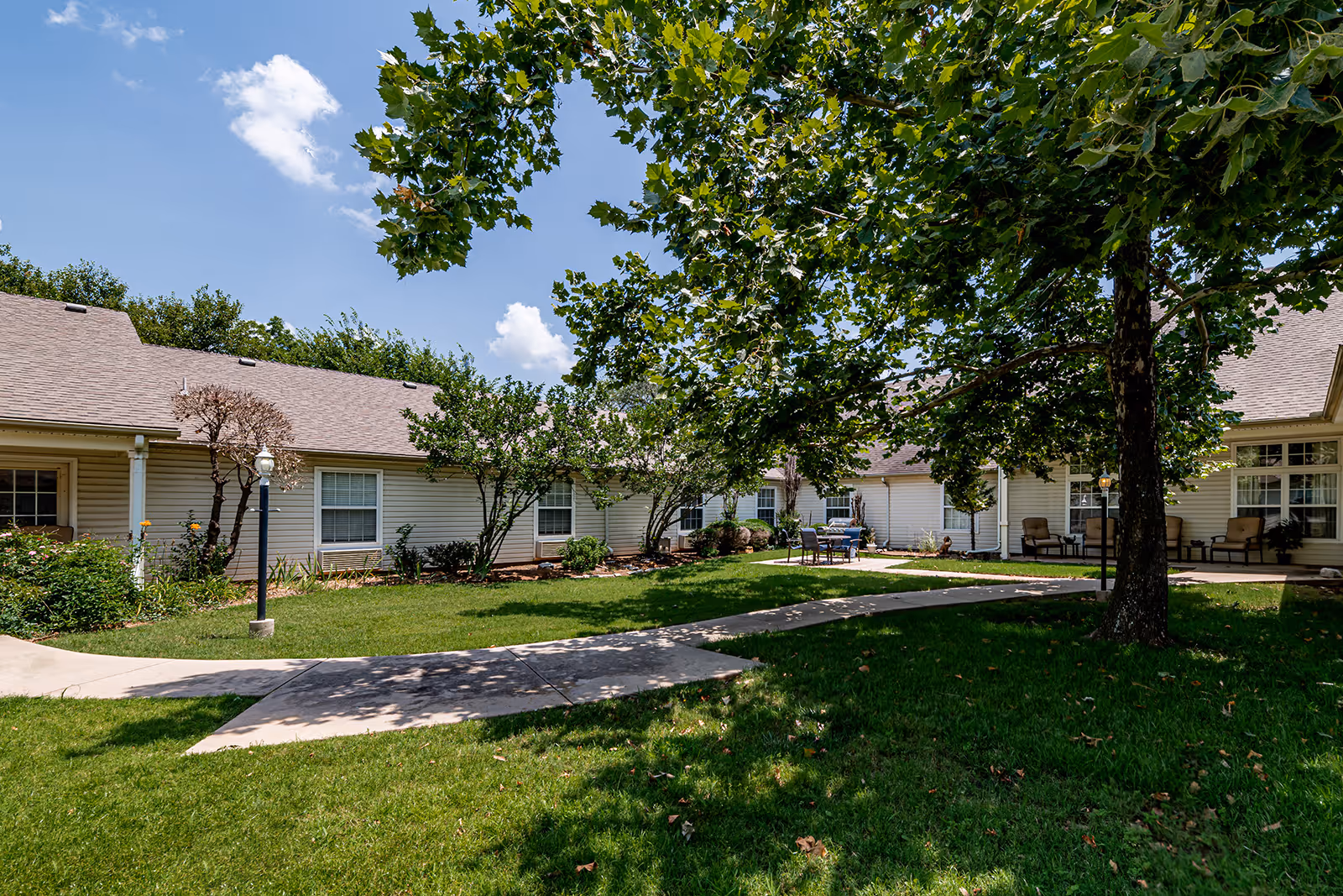 Sunny courtyard with a paved walkway, lawn, trees, and a single-story building with patio seating.