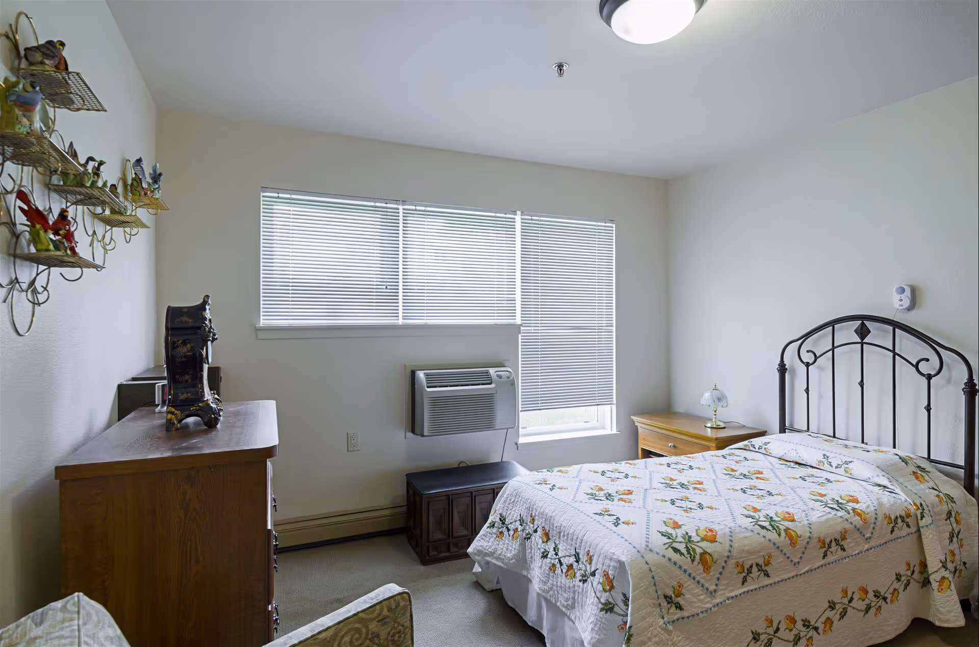 A bedroom with a single bed covered in a white quilt with floral patterns. The room has a metal headboard, a wooden nightstand with a small lamp, a wooden dresser with decorative items on top, and a window with blinds partially closed. There is also a wall-mounted air conditioning unit below the window.