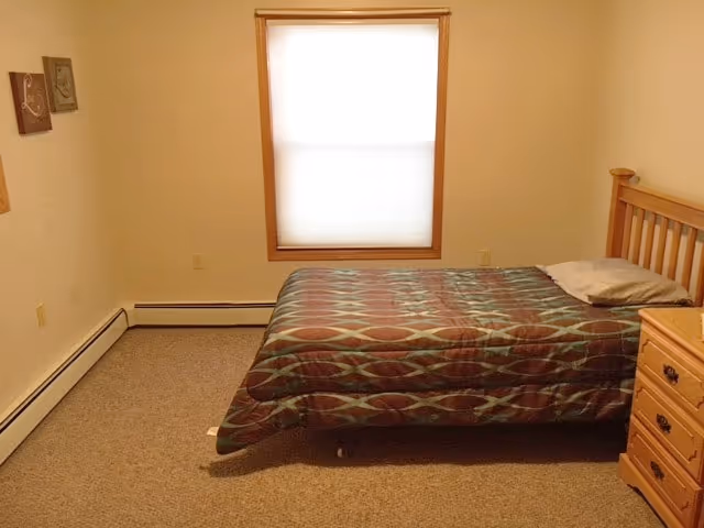 A simple bedroom with a single bed covered with a patterned bedspread, a pillow, a wooden headboard, a wooden nightstand with drawers, a window with a closed blind, and two small framed pictures on the wall.