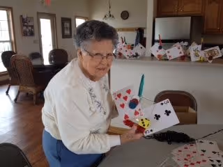 An elderly woman with short gray hair and glasses is sitting at a table in a well-lit room, holding a large fan of oversized playing cards. The room has wooden furniture, including chairs and a table, and there are more oversized playing cards scattered on the table. The background shows windows and a door letting in natural light.