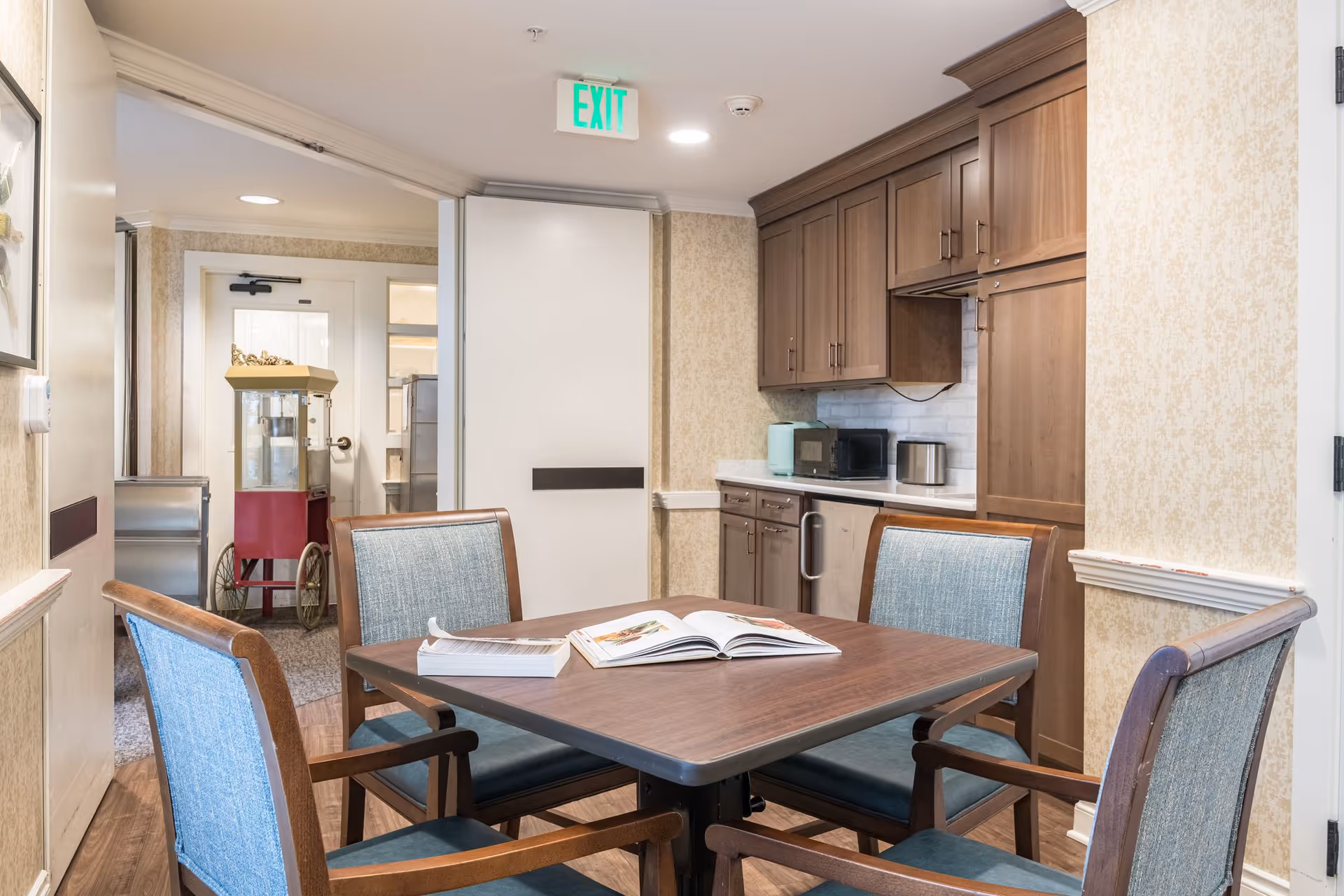 A small dining area in a senior living facility with a square wooden table surrounded by four wooden chairs with blue upholstery. On the table are two open books. Behind the table is a kitchenette with wooden cabinets, a microwave, a toaster, and a small appliance. In the background, there is a hallway with a red popcorn machine and an exit sign above a door.