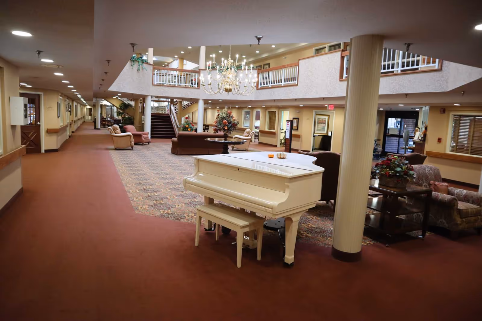 Interior view of a senior living facility lobby area with a white grand piano and bench in the foreground. The space features a large chandelier hanging from the ceiling, comfortable seating arrangements with armchairs and sofas, floral decorations on tables, and a staircase leading to an upper level with railings. The area is carpeted and well-lit with ceiling lights.