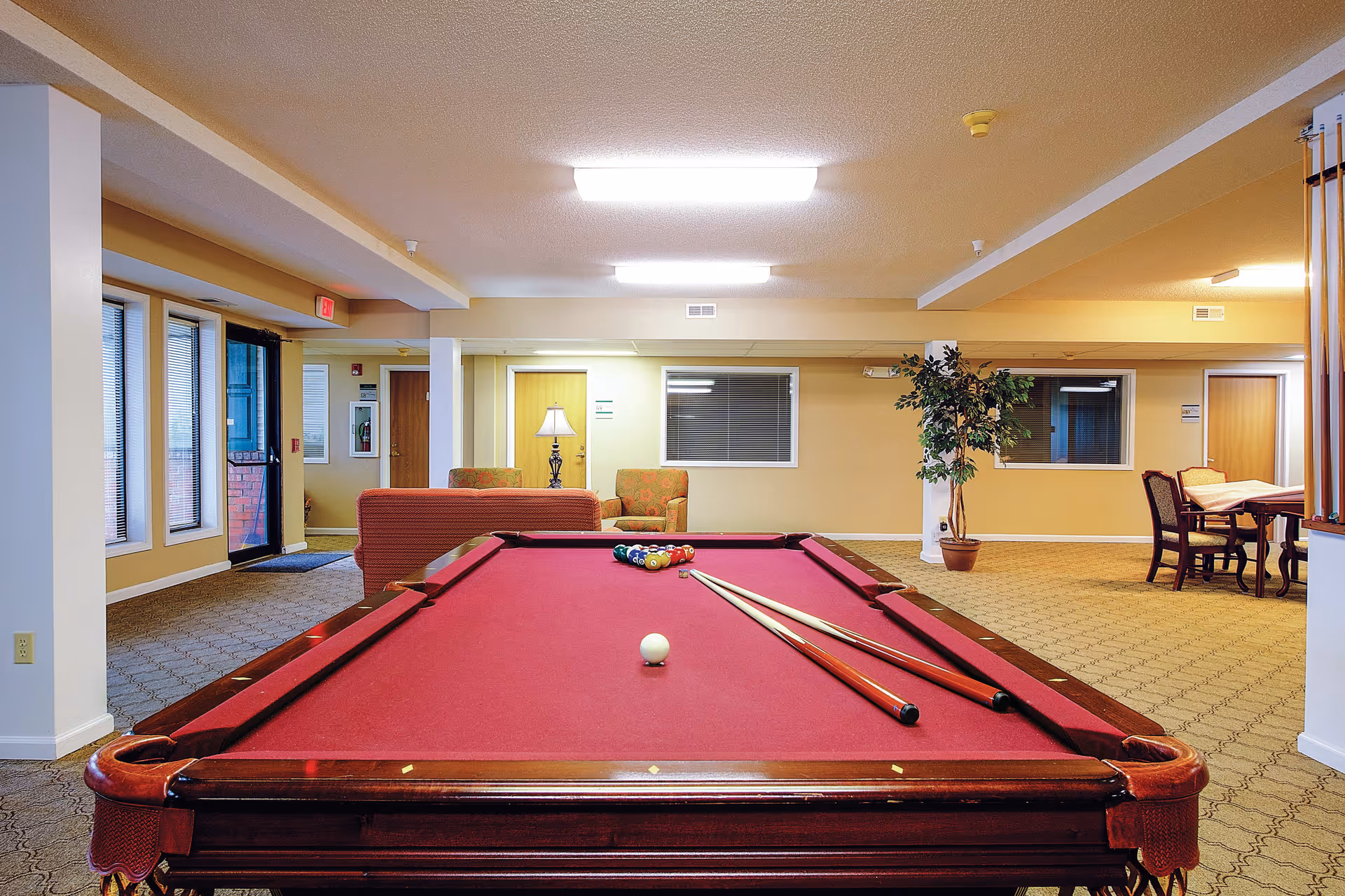 Interior view of a senior living facility common area featuring a red pool table with two cues and a set of billiard balls arranged for a game. In the background, there are comfortable seating areas with armchairs, a lamp, a potted plant, and a table with chairs. The room has beige walls, carpeted floors, and fluorescent ceiling lights.