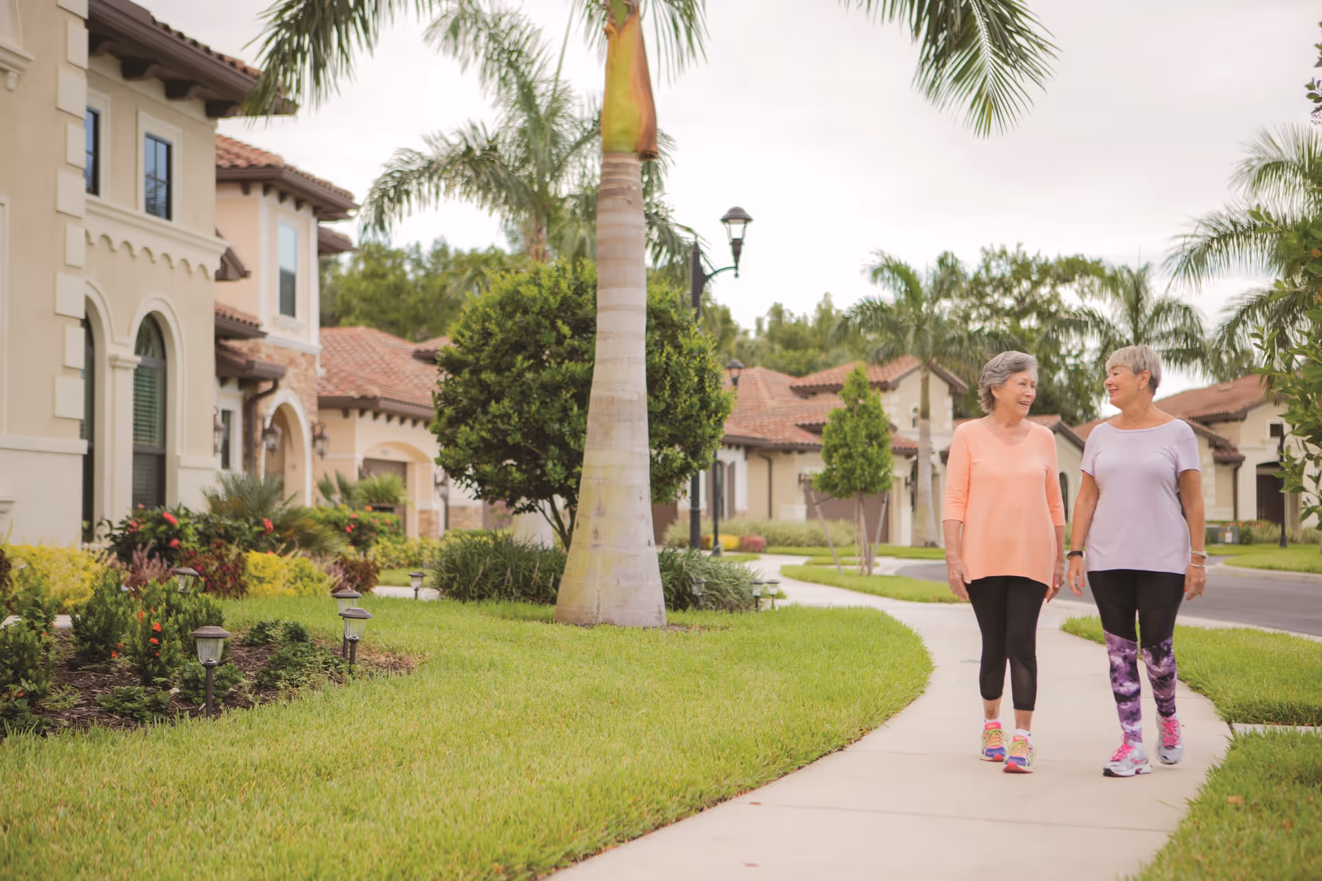 Two elderly women walking and talking on a sidewalk in a residential neighborhood with well-maintained lawns, palm trees, and Mediterranean-style houses in the background.