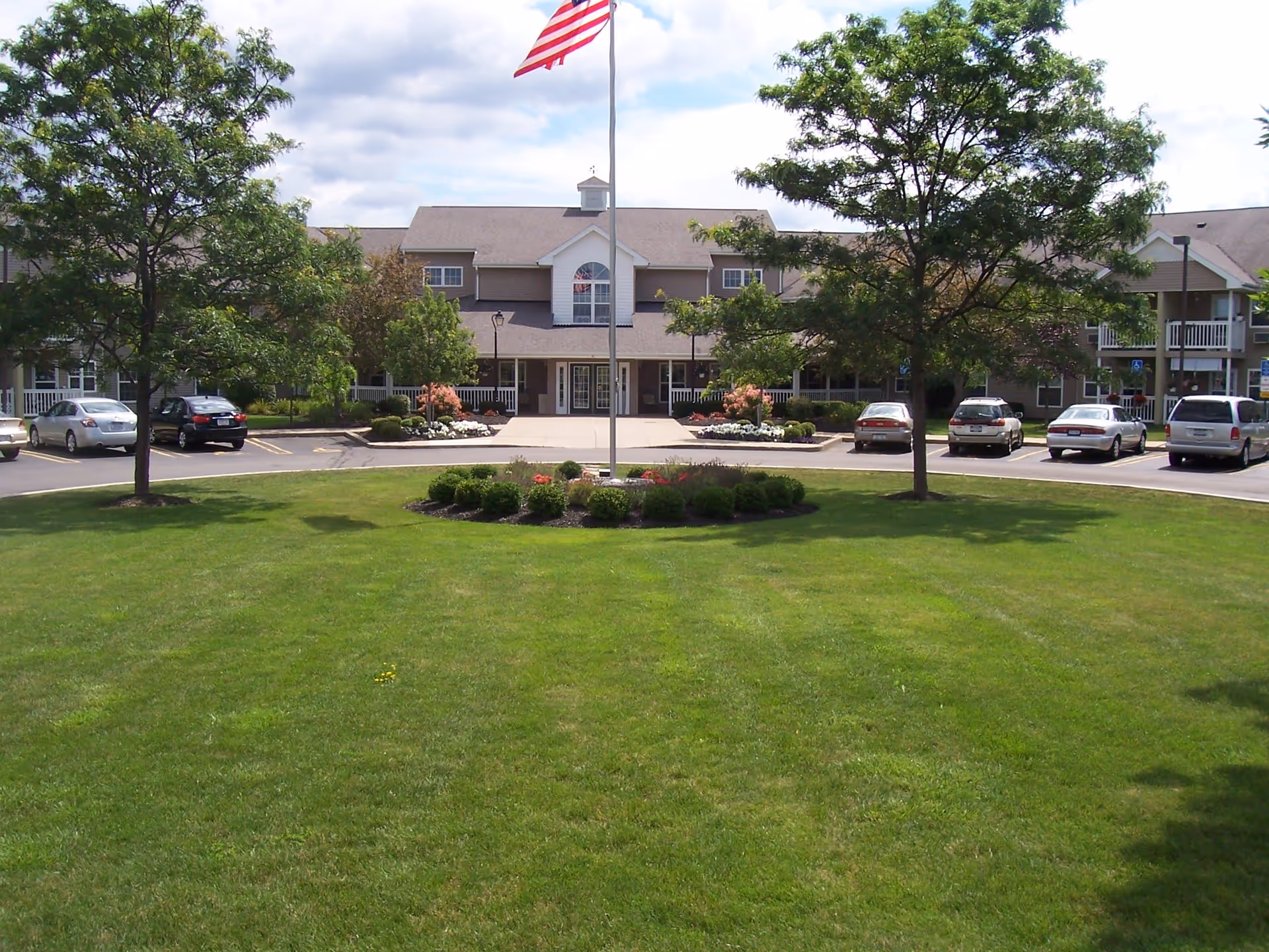 Front exterior view of College Greene Senior Apartments building with a large green lawn, trees, a flagpole with an American flag, and a parking area with several cars.