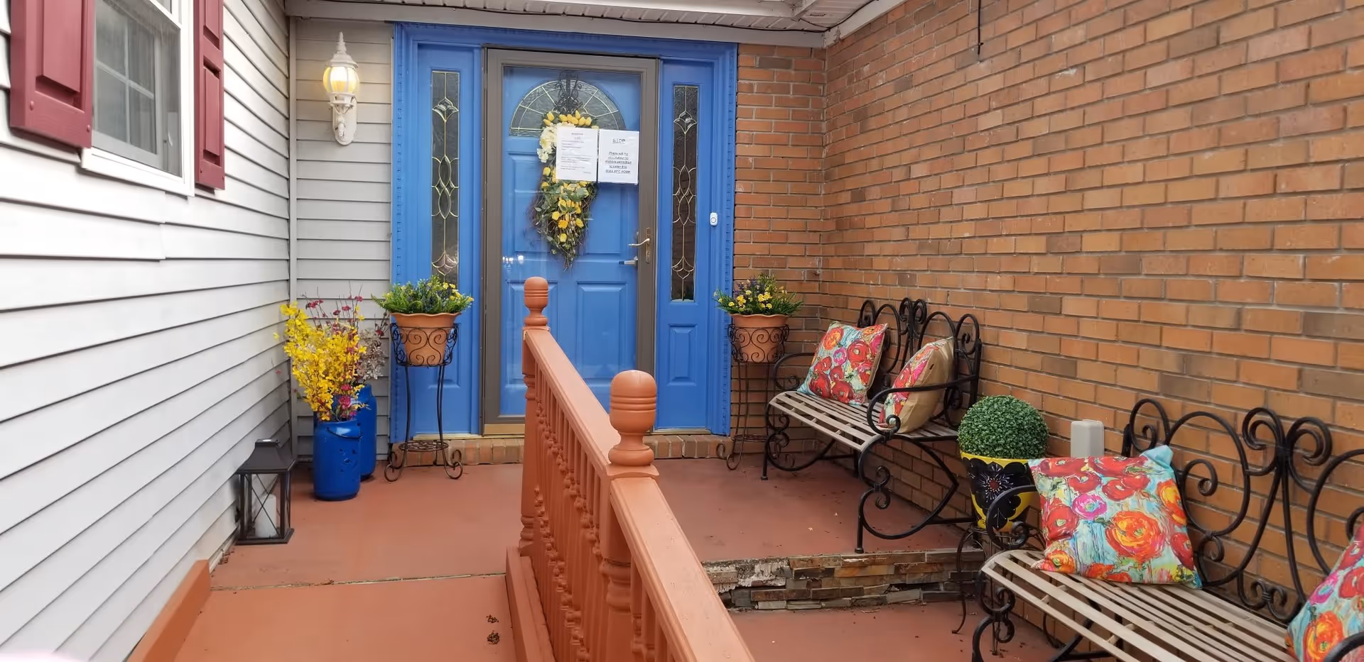 Entrance area with a bright blue door decorated with a yellow floral wreath and two notices attached. The porch has a reddish-brown floor and railing, with two black metal benches adorned with colorful floral cushions. There are potted plants and flowers placed around the entrance, including yellow and purple flowers in blue and terracotta pots. The walls are a combination of white siding with red shutters and red brick.