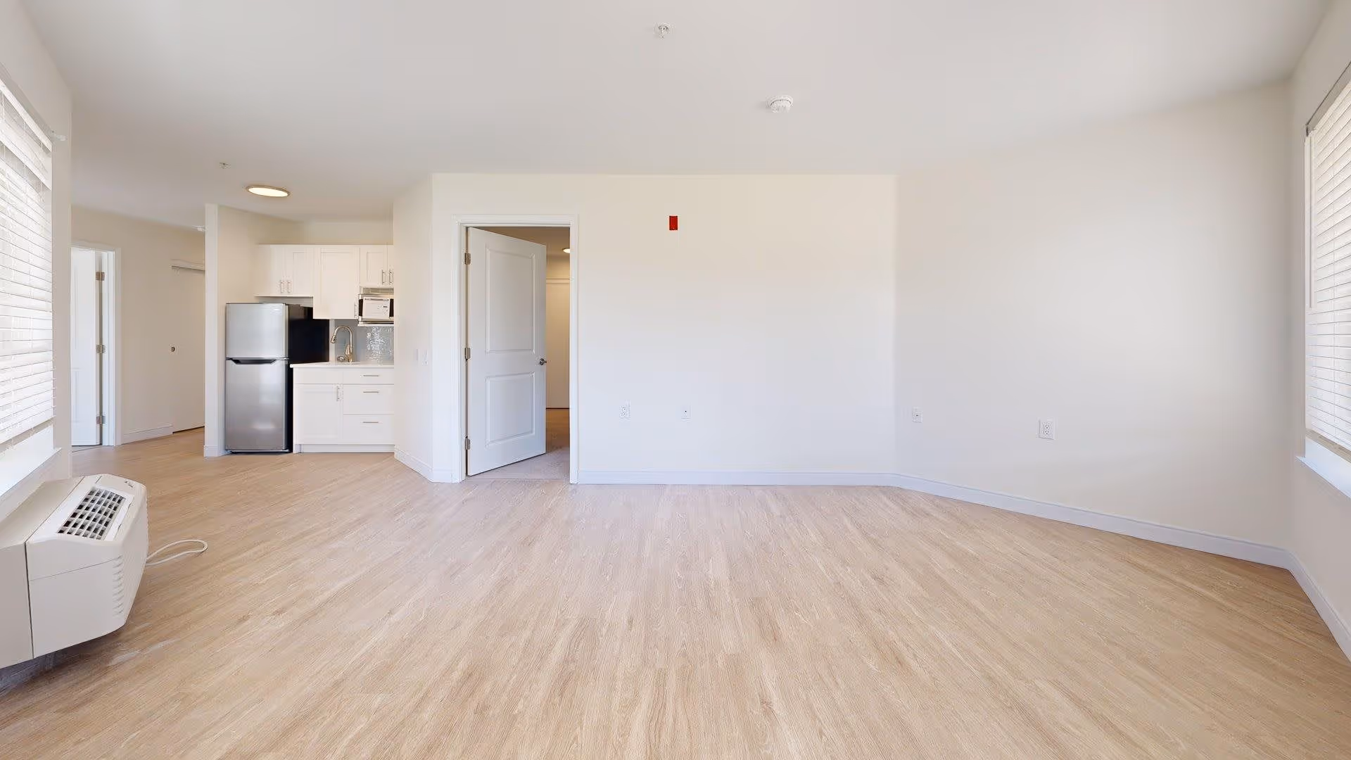 Empty room with light wood flooring, white walls, and two windows with blinds. A small kitchen area with white cabinets, a refrigerator, and a microwave is visible in the background. An open door leads to another room.