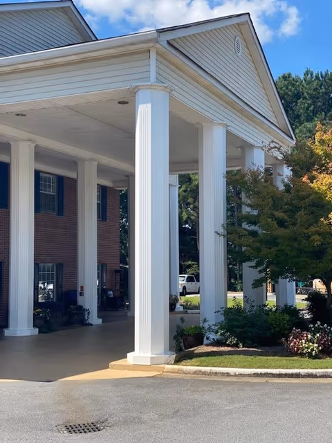 Entrance of a senior living facility with large white columns supporting a covered driveway. The building has red brick walls and several windows with dark shutters. There are some plants and trees near the entrance, and a clear blue sky is visible above.