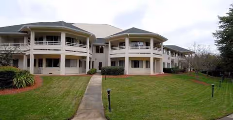 Two-story beige senior living building with balconies, a central walkway, and a manicured lawn.