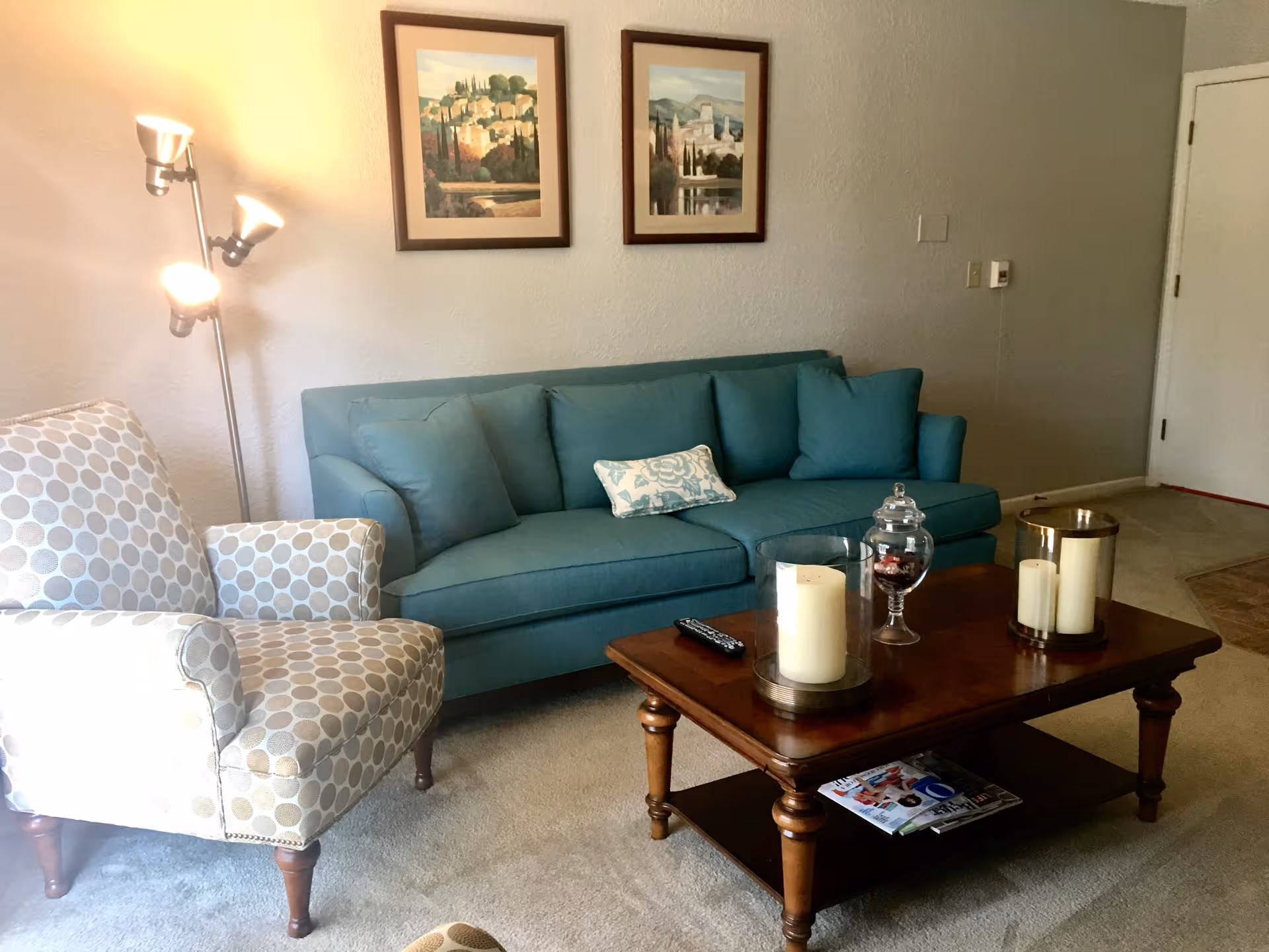 Cozy living room with a teal sofa, patterned armchair, wooden coffee table with candles and decor, and framed wall art.