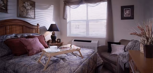 A cozy bedroom with a wooden bed frame, patterned bedding, and several pillows. A wooden tray with a cup and saucer is placed on the bed. Next to the bed is a nightstand with a lamp and decorative items. A window with blinds and sheer curtains lets in natural light. There is a comfortable armchair with cushions and a throw blanket, and a dresser with a vase of dried flowers.