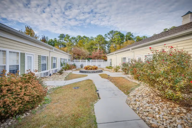 Outdoor courtyard area between two single-story buildings with beige siding and green shutters, featuring a concrete walkway, landscaped bushes, and a circular stone planter with plants in the center under a partly cloudy sky.