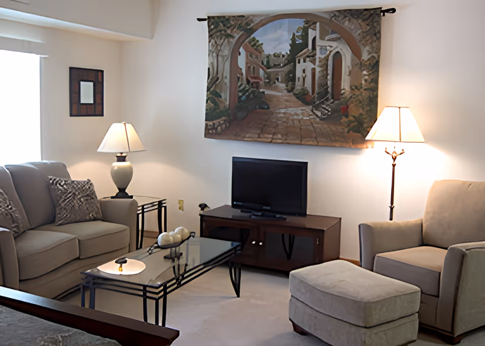 Living room with beige sofa and armchair, glass coffee table, TV on a stand, and a decorative tapestry on the wall.