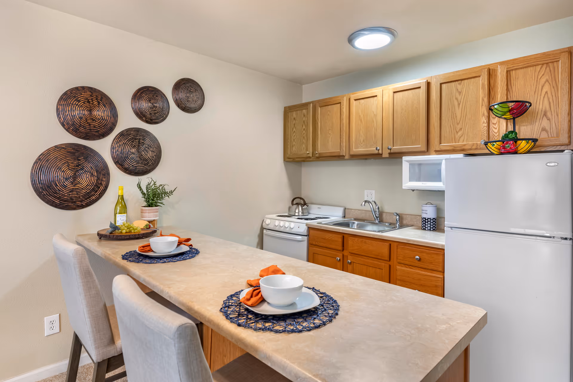 A compact kitchen area with wooden cabinets, a white refrigerator, a small stove, and a sink. A countertop extends outward with two place settings, each with a white bowl, plate, and orange napkin on a blue placemat. Decorative round wall hangings are on the wall, and a bottle of wine with a small plant and grapes are on the counter.