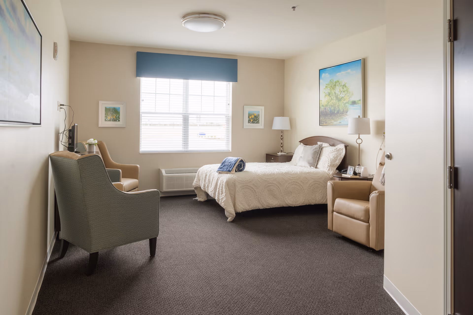 A cozy bedroom in a senior living facility with a bed covered in a white quilt, two armchairs, a window with blinds and a blue valance, two framed pictures on the walls, a nightstand with a lamp, and a painting of a tree above the bed.