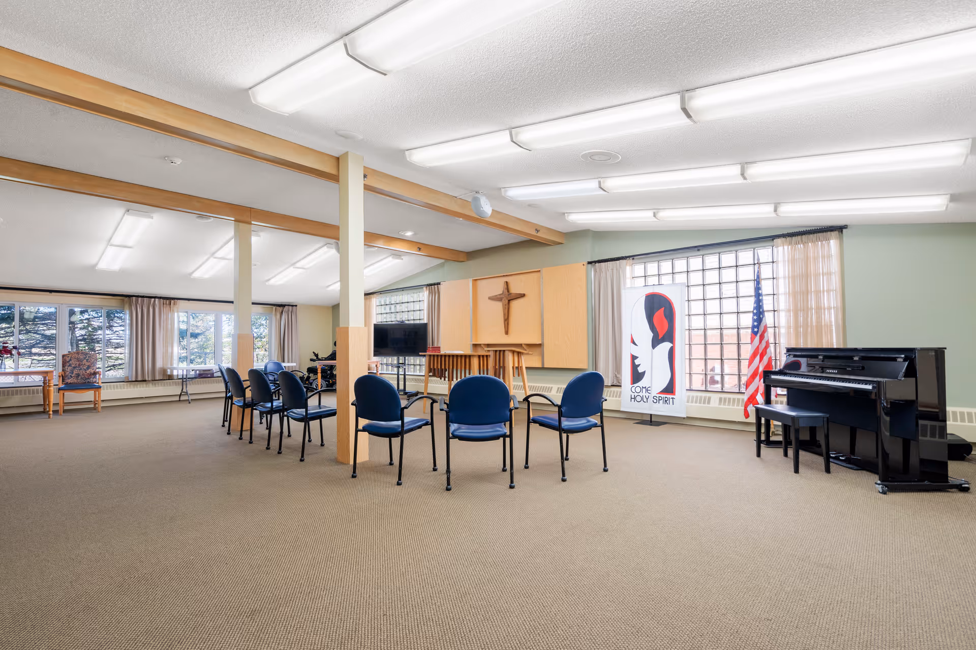 A spacious room with beige carpet and light green walls, featuring a semi-circle of blue chairs facing a wooden podium with a cross mounted on the wall behind it. To the right, there is a black piano with a bench, an American flag, and a banner with a dove and the words 'COME HOLY SPIRIT'. Large windows with curtains allow natural light into the room.